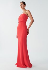 Punainen olkaimeton mekko, jossa on kiinteä vartalo ja levenevä helma. Sujuva tekstuuri, minimalistinen muotoilu ja sulava siluetti korostavat kaaria.