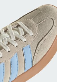 Gros plan d'une basket en daim beige avec des lacets blancs, des bandes latérales bleu clair et une semelle en caoutchouc naturel.