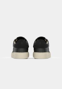 Vue arrière de baskets basses noires avec des semelles blanches épaisses sur un fond gris clair uni.