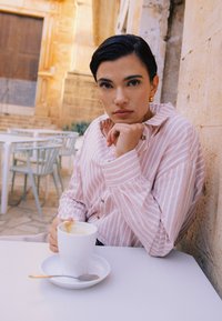 Personne aux cheveux courts et foncés portant une chemise rayée rose, assise à une table de café en plein air, tenant une tasse de café et regardant la caméra.