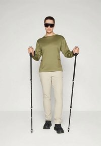 Olivgrön långärmad skjorta, beige byxor, svarta mobilitetsstavar i händerna och svarta skor. Modellen bär solglasögon och står upprätt.