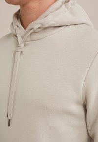 Svetlo bež kapuca z mehko teksturo, okrašena z vrvico za nastavitev in sproščenim krojem. Tkanina deluje gladko z subtilnimi detajli.
