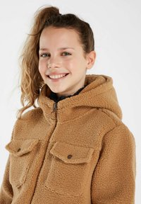 Giacca in pile beige con cappuccio, dotata di due tasche sul petto, chiusure a scatto e un tessuto testurizzato. Chiusura frontale con zip e cursore rotondo.
