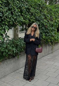Stort sort blazer over en gennemsigtig sort blonde maxiskirt. Accessories inkluderer en lille burgunder taske og mørke solbriller. Grønne planter i baggrunden.