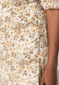 Robe à motif floral dans des tons crème, vert clair et marron. Dispose d'une poche latérale et de détails à boutons le long de la couture. Le tissu présente une texture douce.