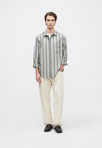 Camisa de botones a rayas gris y blanca con mangas enrolladas, bolsillo frontal, combinada con pantalones beige claros y zapatos oscuros.