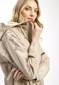 Beżowa wodoodporna kurtka z kapturem; posiada elastyczne mankiety, detale na zatrzaski oraz gładką, błyszczącą teksturę.