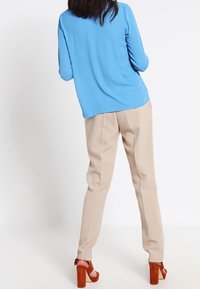 Femme debout, vue de dos, portant un chemisier bleu à manches longues, un pantalon beige et des sandales marron à talons hauts, sur un fond blanc.