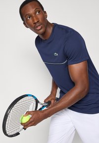 Granatowy sportowy t-shirt z kontrastowymi białymi wykończeniami, z zielonym logo. Model trzymający rakietę tenisową i piłkę, z krótkimi rękawami.