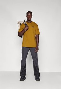 Άνδρας με μουσταρδί μπλουζάκι, σκούρους δίχρωμους παντελόνες και μαύρα παπούτσια, κρατώντας μια μπεζ θήκη σακιδίου πάνω στον ώμο του, στέκεται σε απλό φόντο.