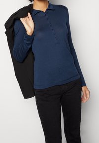 Person klädd i en marinblå långärmad knäppt skjorta och svarta byxor, som håller en svart jacka över axeln.