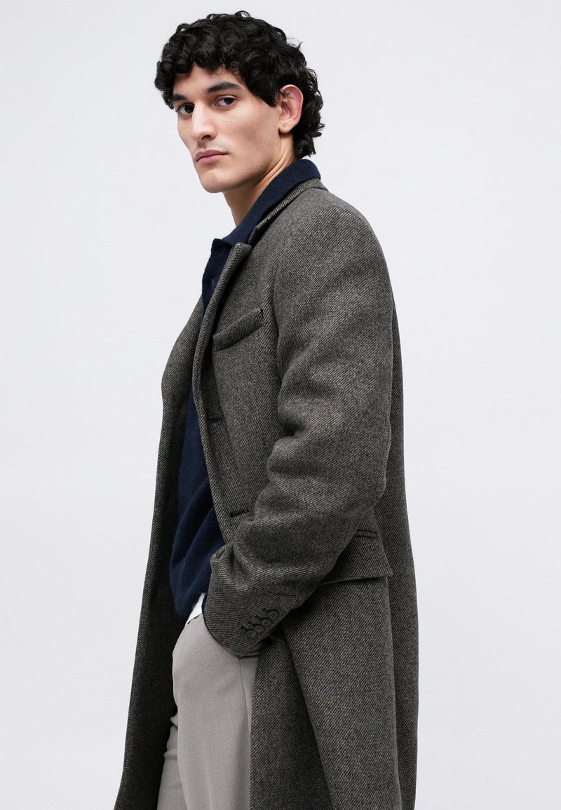 Homme avec des cheveux bruns bouclés portant un pardessus en chevron gris, un pull bleu marine et un pantalon gris clair, se tenant avec une main dans sa poche.