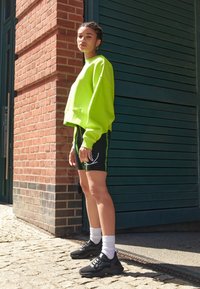 Neon zelená oversized mikina, černé kraťasy se zelenými akcenty, černé tenisky a bílé ponožky na pozadí cihlové zdi a zelených dveří.