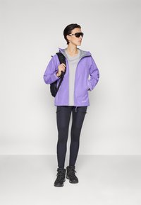 Lilla vanntett jakke med grå innside, kombinert med en grå skjorte, sorte leggings og sorte tursko. Svart ryggsekk og solbriller inkludert.