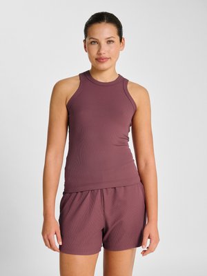 Młoda kobieta z ciemnymi włosami związanymi do tyłu, ubrana w bezrękawnik bordowy i pasujące krótkie spodenki, stoi na jednolitym jasnym tle.