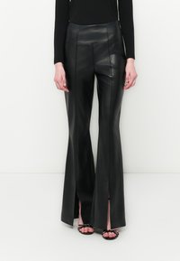 Pantaloni finti in pelle neri a vita alta svasati con fessure frontali, indossati con un top nero a maniche lunghe e tacchi aperti neri con cinturini.