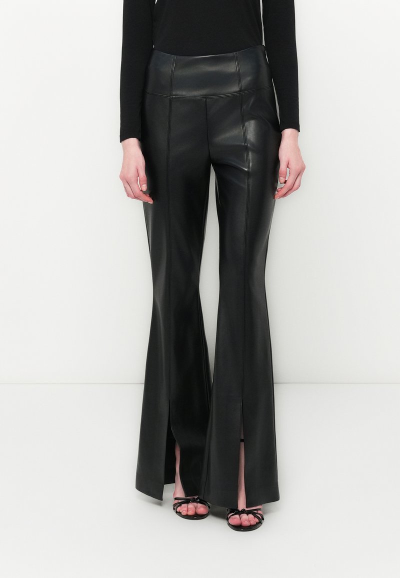 Pantaloni finti in pelle neri a vita alta svasati con fessure frontali, indossati con un top nero a maniche lunghe e tacchi aperti neri con cinturini.