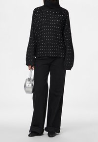 Svart stickad polotröja med silvernitar, tillsammans med vida svarta byxor. Håller en liten silverhandväska med rundad form.