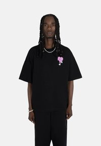 Svart oversized t-skjorte laget av bomull, med et rosa hjertegrafikk på brystet. Korte ermer og en avslappet passform.