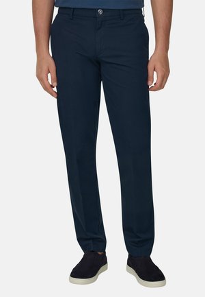 Homme portant un pantalon droit bleu foncé et des chaussures noires à enfiler avec des semelles blanches, debout devant un fond uni.
