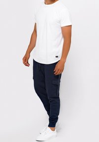 Mężczyzna w prostym białym t-shircie z krótkim rękawem, granatowych spodniach dresowych cargo i białych trampkach stoi na jasnym tle.