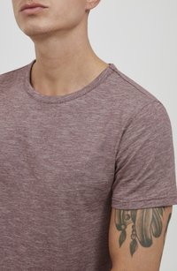 Mørk rød heathered t-skjorte med rund hals, korte ermer, og en myk tekstur; synlig tatovering på venstre arm.