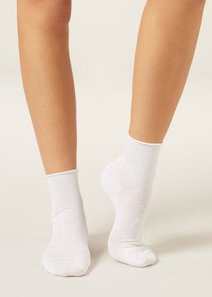 Nohy v jednoduchých bílých kotníkových ponožkách stojí na světlém povrchu, jedna noha je položena celou plochou a druhá na špičce.