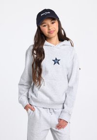 Lichtgrijze hoodie met een buidelzak aan de voorkant en een donkerblauw sterrenpatroon. Gecombineerd met bijpassende sweatpants met trekkoord. Zwarte pet met de tekst "New York".