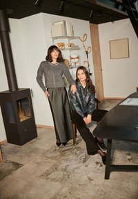 Dos mujeres en un interior moderno. Una lleva un cardigan gris sobre una falda larga de satén oscuro; la otra viste una camisa de cuero negra y pantalones.