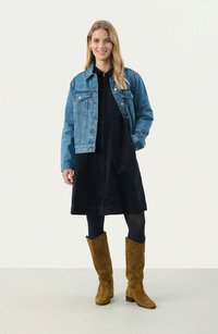 Giacca di jeans blu chiaro, vestito in corduroy nero, stivali al ginocchio in suede marrone. Modella in piedi, mani nelle tasche della giacca, sfondo neutro.
