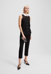 Svart ermeløs bluse med hvit krage og gullknapper, kombinert med svarte capribukser og svarte spisse hæler, holder en svart clutch.