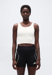 Αθλητική γυναίκα όρθια, φοράει λευκό κοντό τοπ και στενά μαύρα σορτς με λευκό περιθώριο, σε απλό λευκό φόντο.