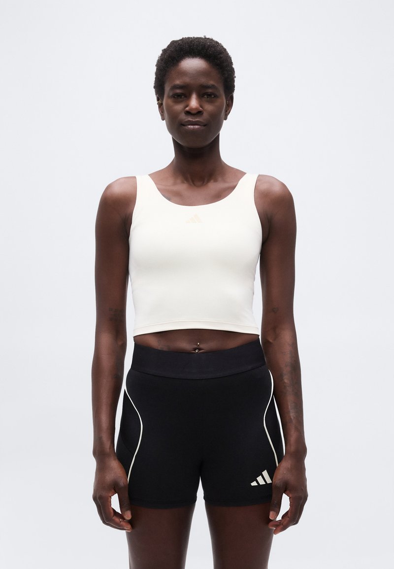 Αθλητική γυναίκα όρθια, φοράει λευκό κοντό τοπ και στενά μαύρα σορτς με λευκό περιθώριο, σε απλό λευκό φόντο.