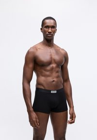 Bóxer negro hecho de una tela suave, con un ajuste ceñido y una banda con logo en la cintura, usado por un modelo masculino de complexión atlética.