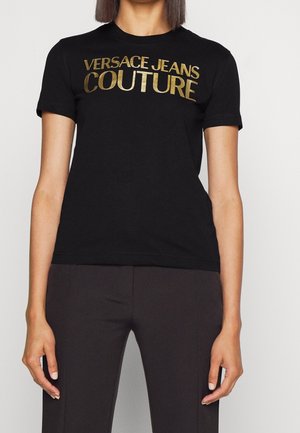 Femme portant un t-shirt noir à manches courtes avec un texte doré "Versace Jeans Couture" et un pantalon ajusté foncé sur un fond uni.