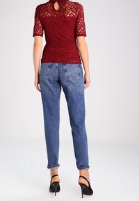 Burgunderfarbener Spitzenoberteil mit kurzen Ärmeln, Rückendetail mit Schlüsselloch, kombiniert mit hellblauen lockeren Jeans und schwarzen High-Heels.