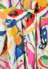Tissu vibrant avec des motifs floraux et fruitiers abstraits en rouge, jaune, bleu, rose, vert et blanc, avec des boutons marron et une ceinture nouée.
