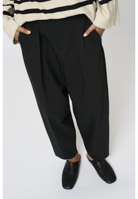 Mains dans les poches d’un pantalon noir ample associées à des chaussures noires à bout carré et un pull crème à rayures noires.