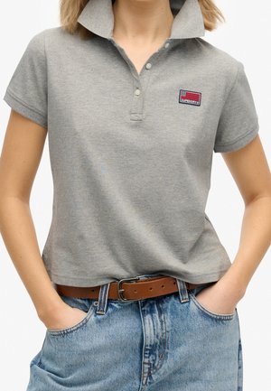 Graues Polohemd mit kurzen Ärmeln und aufgestelltem Kragen, mit einem in Rot und Blau gestickten Logo. Lässig getragen zu hochgeschnittenen Jeans.