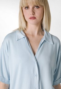 Camisa de botones de color azul claro, hecha de una tela suave, con cuello, mangas cortas y un corte relajado. Los botones son de un azul a juego.