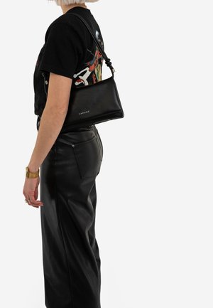 Personne portant un t-shirt graphique noir et un pantalon en cuir noir, portant un sac à bandoulière en cuir noir avec le logo "EXPATRIE".