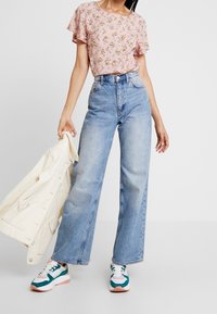 Ljusrosa blus med blommönster och volangärmar, höga vida blå jeans och flerfärgade sneakers. Håller en cremefärgad jacka.