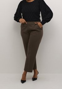 Personne portant un pantalon marron sur mesure, des talons pointus noirs et un chemisier noir à manches longues texturé, debout devant un fond clair uni.