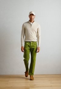 Lys beige zip-neck sweater, grønne kordfløjspants, brune sko og en matchende beige cap; står foran en neutral baggrund.