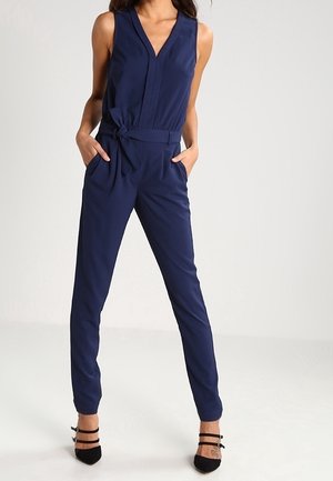 Femme portant une combinaison bleu marine sans manches avec un col en V et une ceinture nouée, debout les mains dans les poches et des talons noirs à brides.