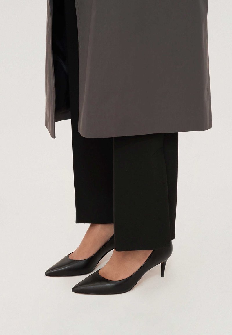 Black leather pointed-toe heeled shoes with a low stiletto heel, worn with black wide-leg trousers and a gray outer garment.