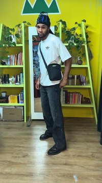 Czarna torebka na ramię, biała koszulka z zielonym logo, duże czarne spodnie, czarne trampki, stoi w pokoju z zielonymi półkami i książkami.