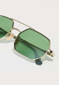 Ochelari de soare hexagonali verzi cu ramă de metal aurie. Caracteristici includ un accent maro deschis pe braț și un logo de marcă vizibil pe una dintre lentile.