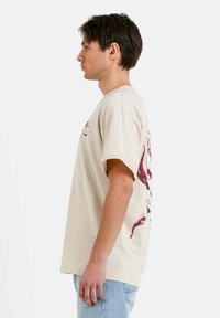 Camiseta de algodón beige con mangas cortas, que presenta una impresión gráfica en burdeos en la parte posterior. Corte estándar, escote redondo y textura suave.