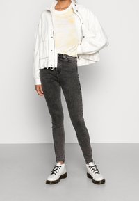 Una persona se encuentra en un entorno minimalista, vistiendo una chaqueta blanca, una camisa amarilla, jeans negros y botas blancas, con una pose de confianza.